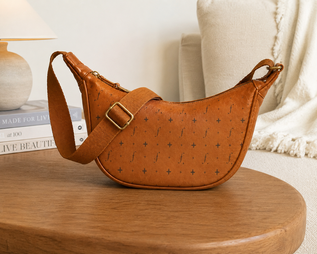 Tan Frounder Luna leather crescent sling bag with brass buckle on a wooden table