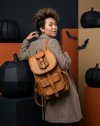 A woman wearing a brown coat and carrying a tan leather backpack with a buckle closure, posed in a Halloween-themed studio setting with black pumpkins and bat decorations.