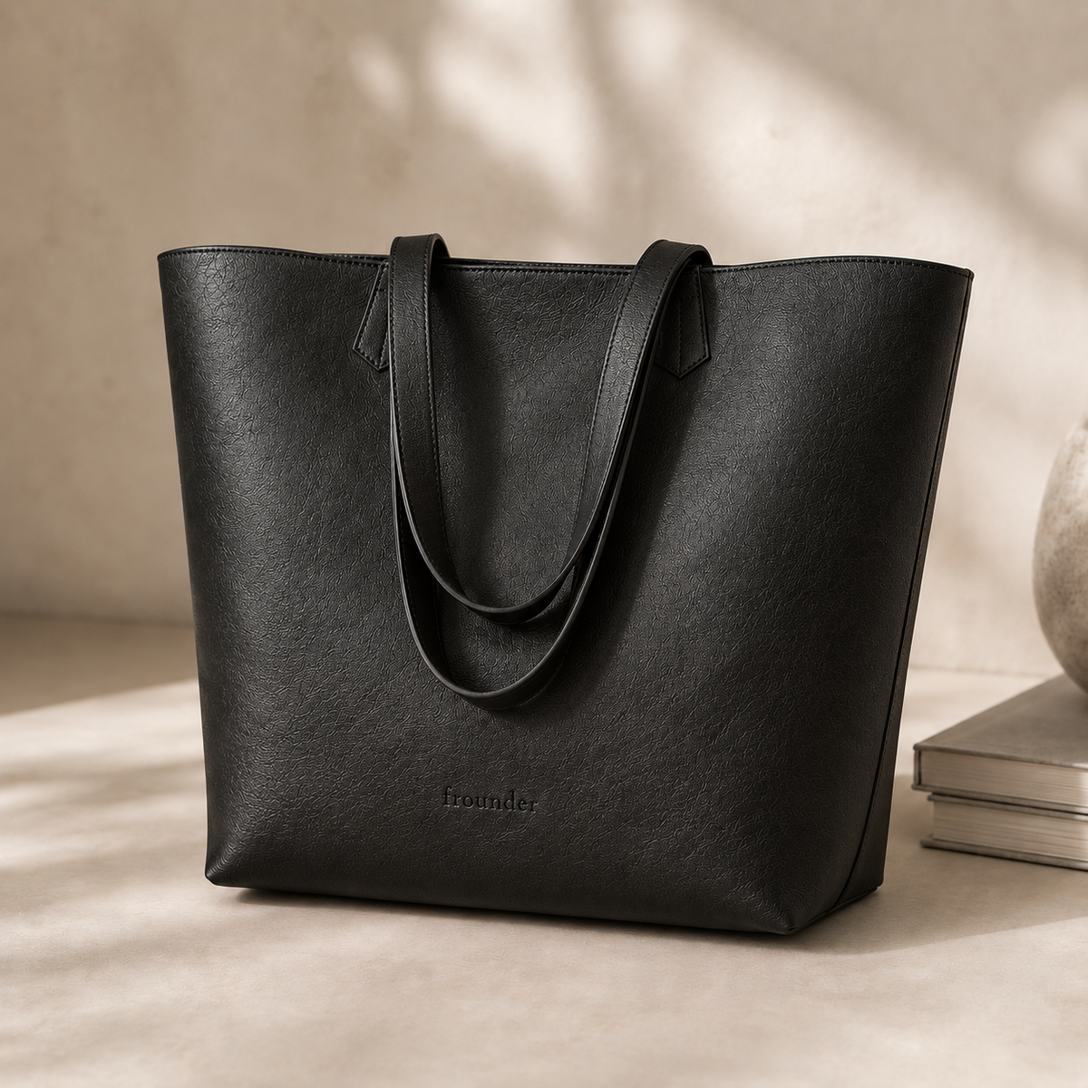 The Altair tote in black, standing upright on a beige surface, showing the double shoulder straps and the embossed Frounder mark low on the front panel.