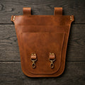 A brown leather utility pouch with two top straps and two front metal snap hooks, laid flat on a dark wood plank background.