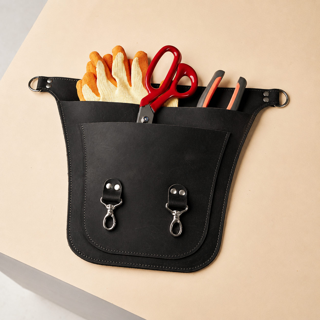 A black leather tool pouch organizer with silver hardware, shown holding work gloves, red-handled scissors, and utility knives on a beige surface.
