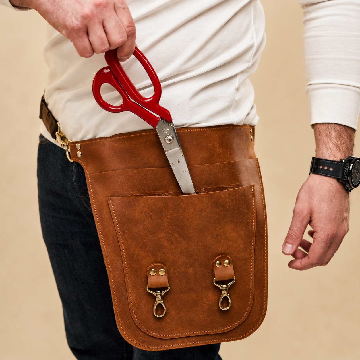 A brown leather tool pouch worn around a person's waist, holding red-handled scissors in its front pocket. The pouch has two brass-tone snap hooks and is shown against a plain beige background.