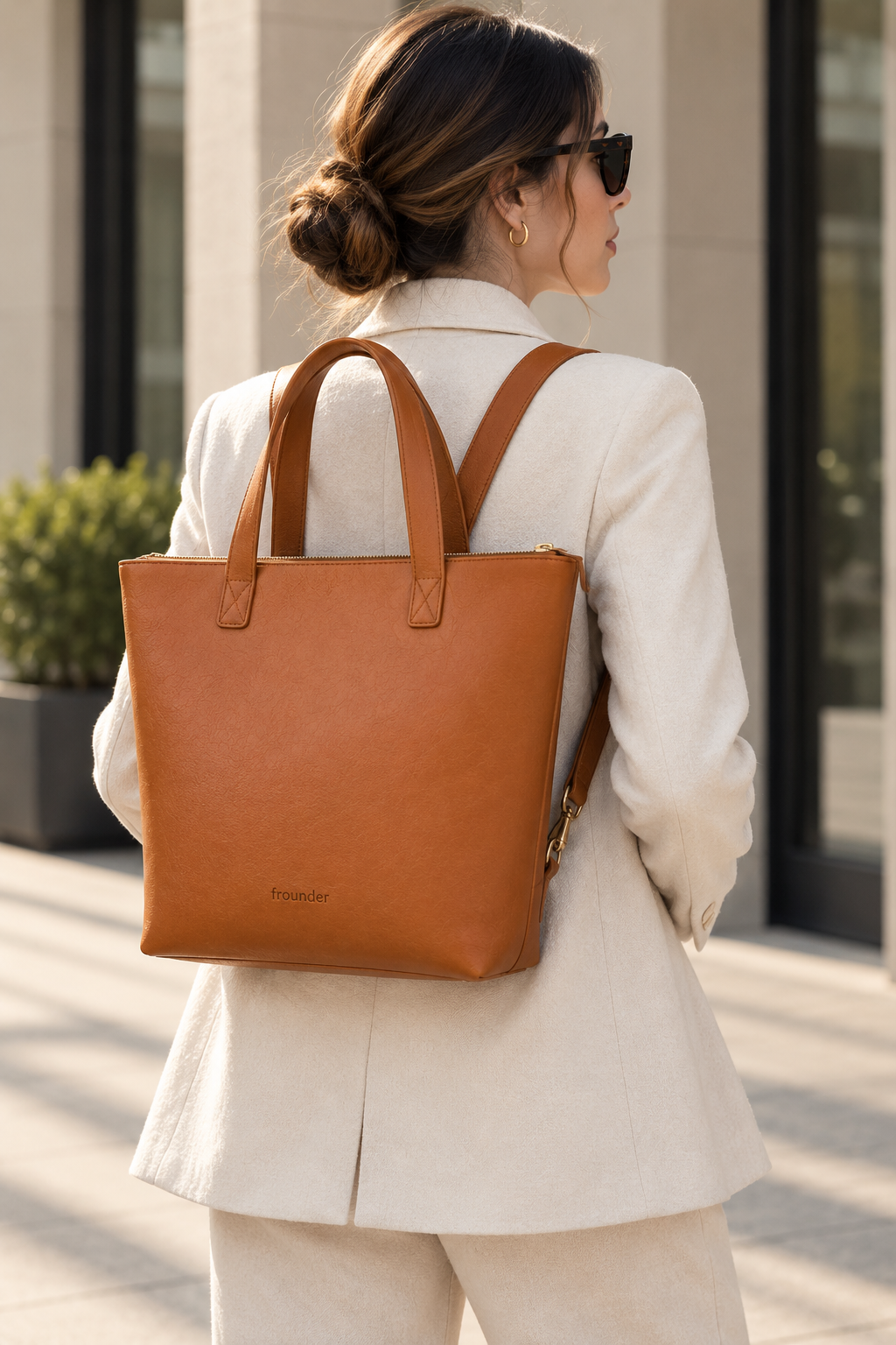 A woman in a white blazer and sunglasses carries a tan leather tote backpack with double top handles in an outdoor urban setting.